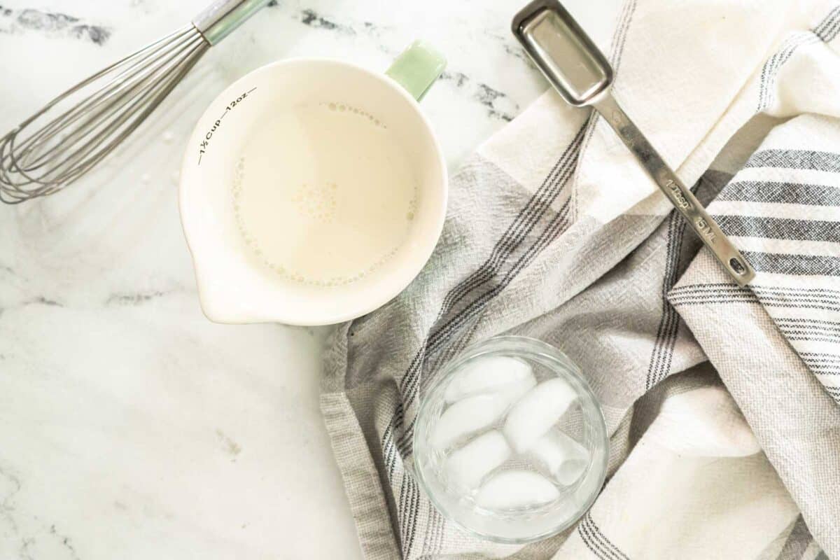 A measuring cup with sour cream, a whisk, a metal tablespoon, a glass of ice water, and a striped kitchen towel on a marble surface.
