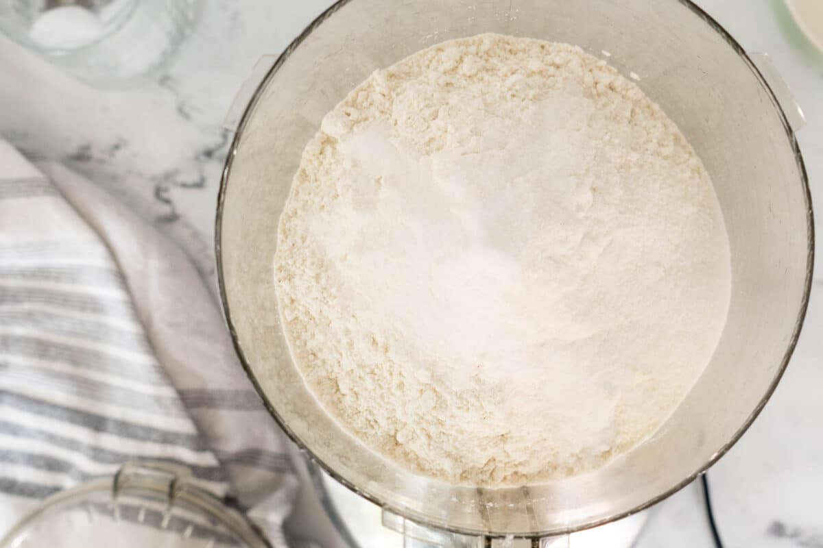 A metal mixing bowl filled with flour sits on a marble countertop next to a striped kitchen towel and a glass container.
