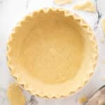 Unbaked pie crust with fluted edges in a white dish, featuring fork marks on the base, sits on a marble surface with dough scraps nearby.