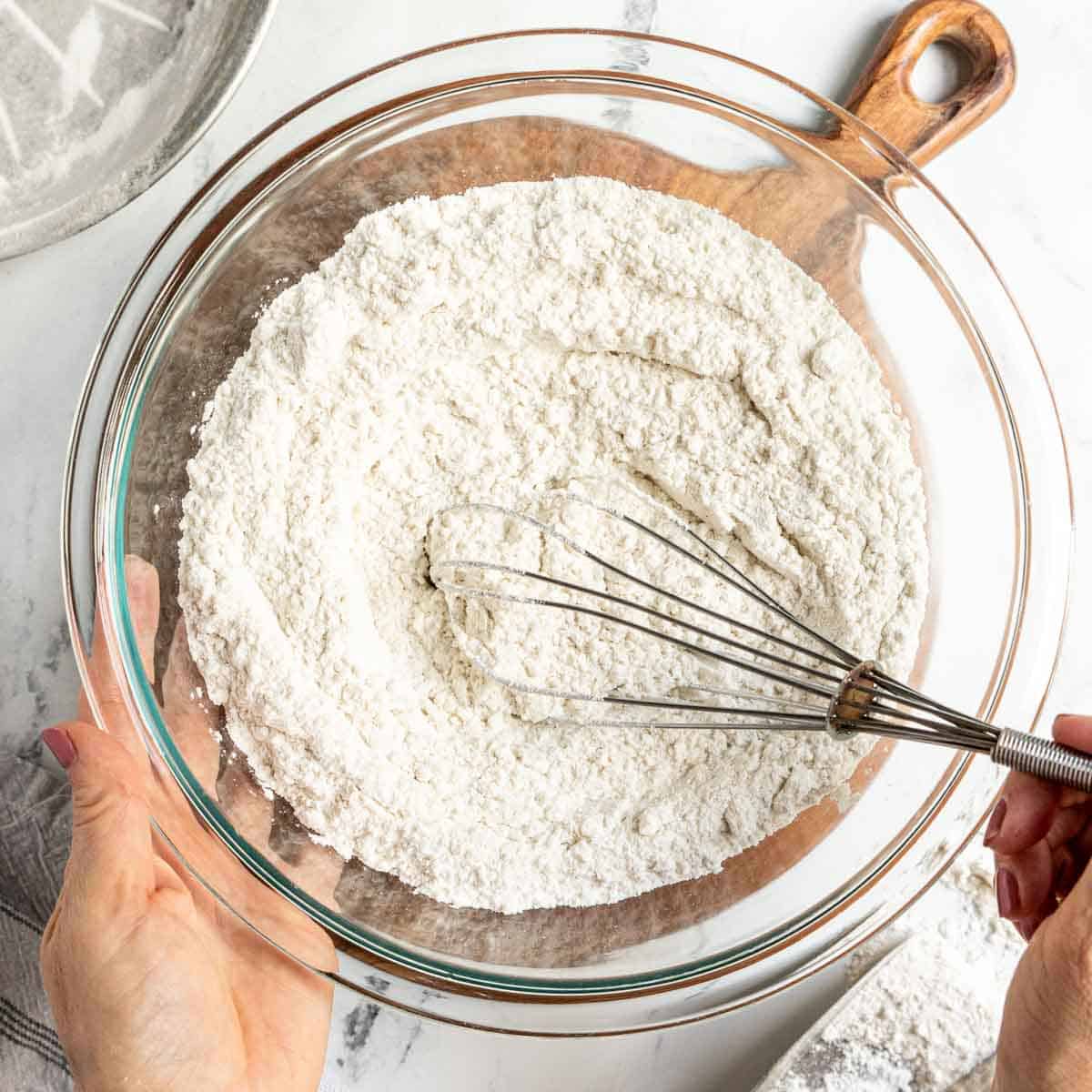 A person whisking gluten free flour in a large glass bowl placed on a wooden board.