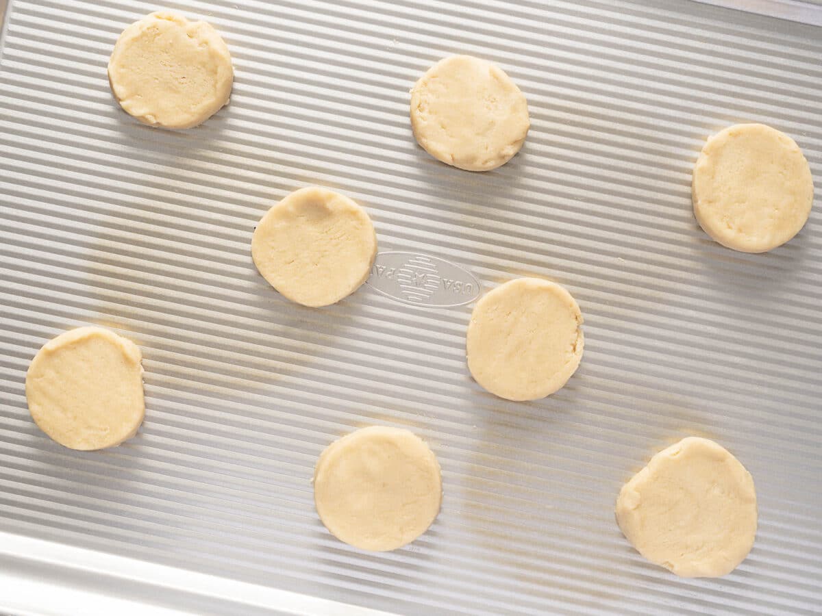 Eight unbaked round cookie dough pieces are spaced out on a silver, ridged baking sheet.