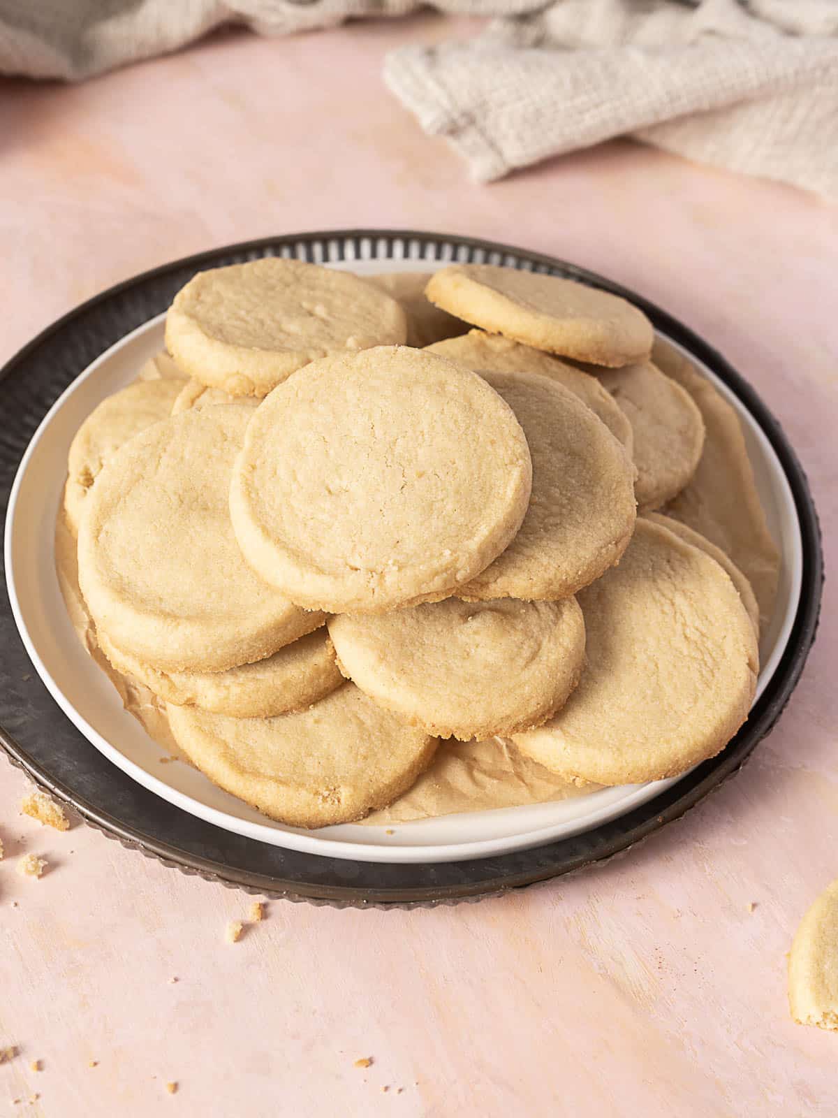 A plate of round, plain shortbread cookies stacked on parchment paper, with a light pink surface and beige cloth in the background.