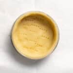 A round, unbaked pie crust in a metal tin with fork marks on the surface, placed on a sheet of white parchment paper.