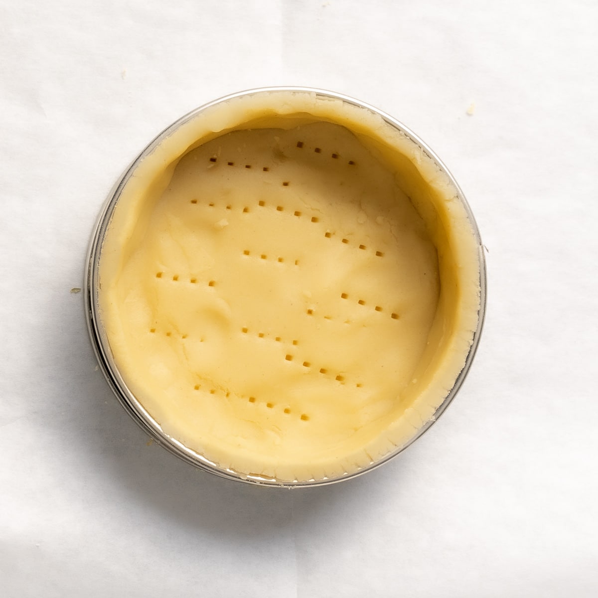 A round, unbaked pie crust in a metal tin with fork marks on the surface, placed on a sheet of white parchment paper.