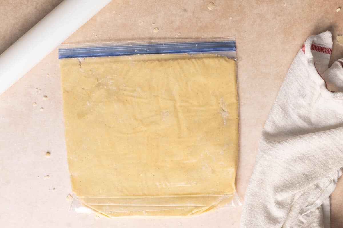 A zip-top plastic bag filled with dough sits on a countertop next to a rolling pin and a folded white kitchen towel.