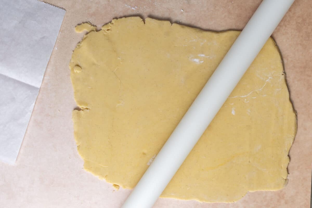A sheet of dough is being rolled out with a white rolling pin on a lightly floured surface, with parchment paper nearby.