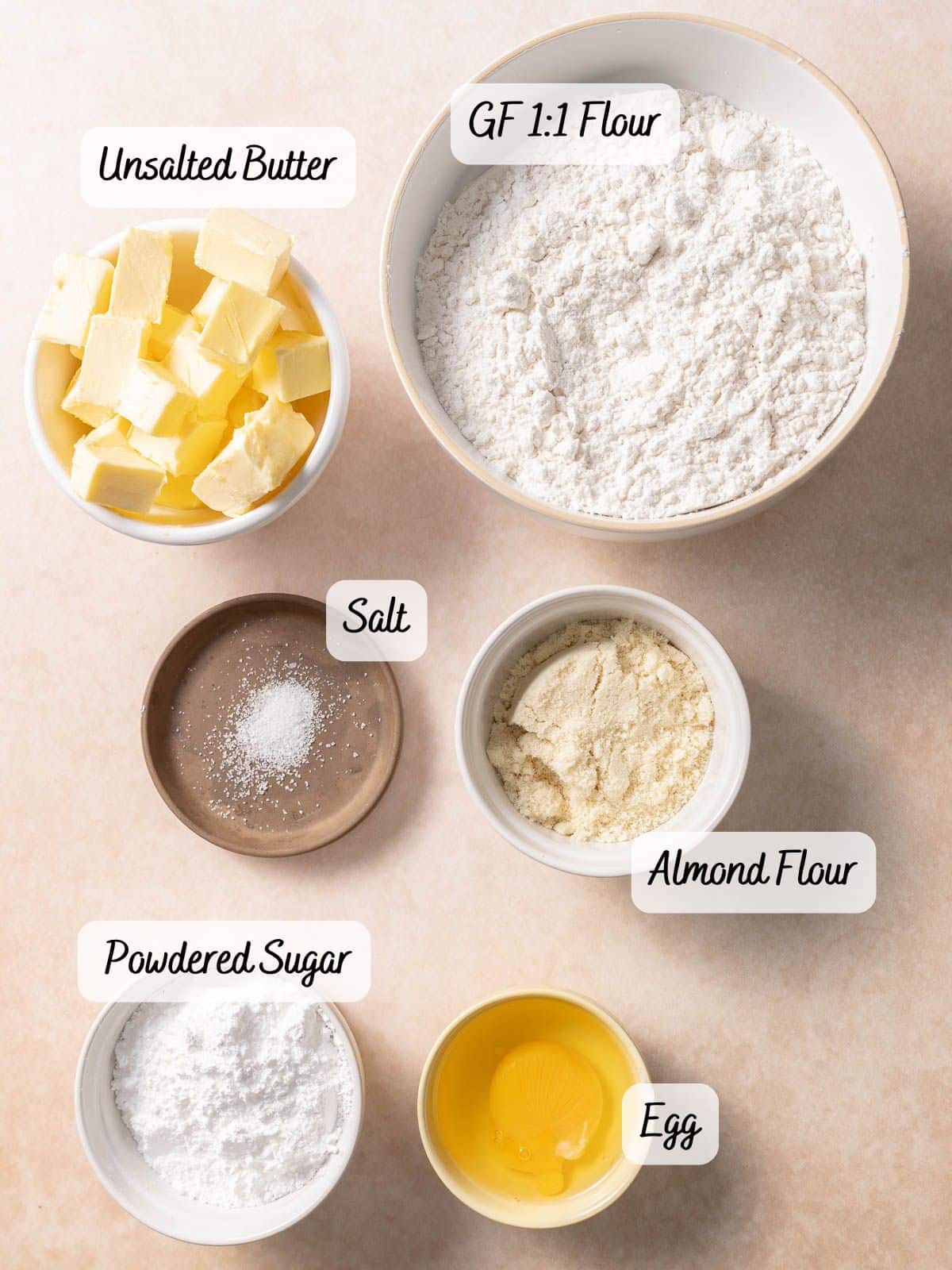 Top-down view of labeled baking ingredients in bowls: unsalted butter, gluten-free 1:1 flour, salt, almond flour, powdered sugar, and a cracked egg.