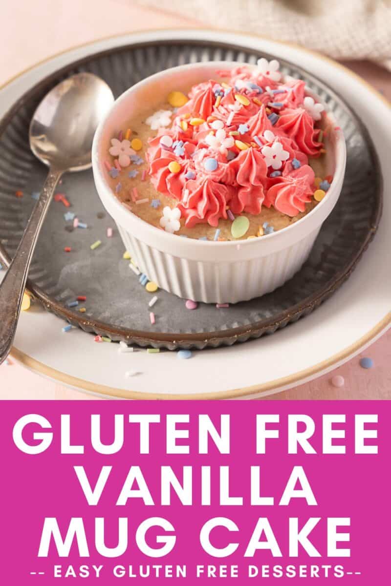 A white ramekin filled with a vanilla mug cake topped with pink frosting and colorful sprinkles sits on a plate with a spoon beside it.