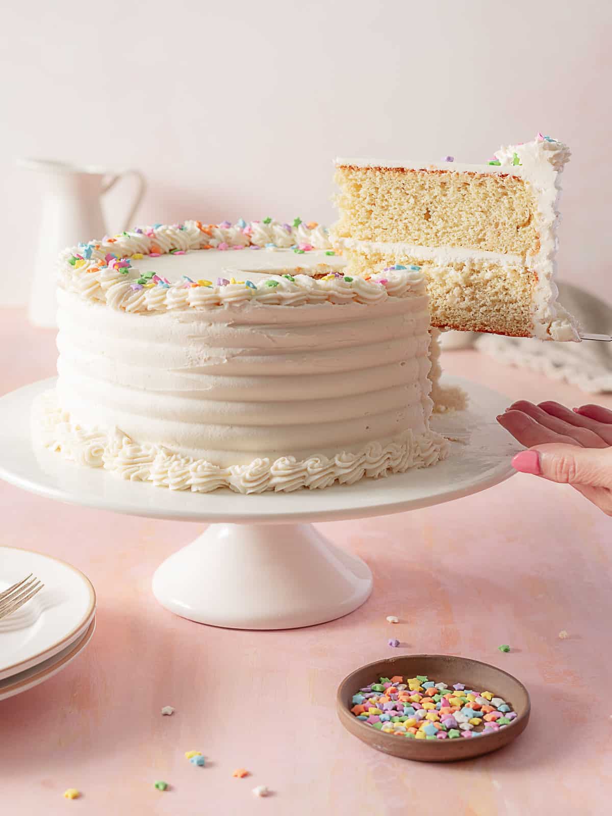 A hand lifts a slice of white-frosted layer cake decorated with colorful star sprinkles from a cake on a white stand. A small bowl of sprinkles sits nearby.