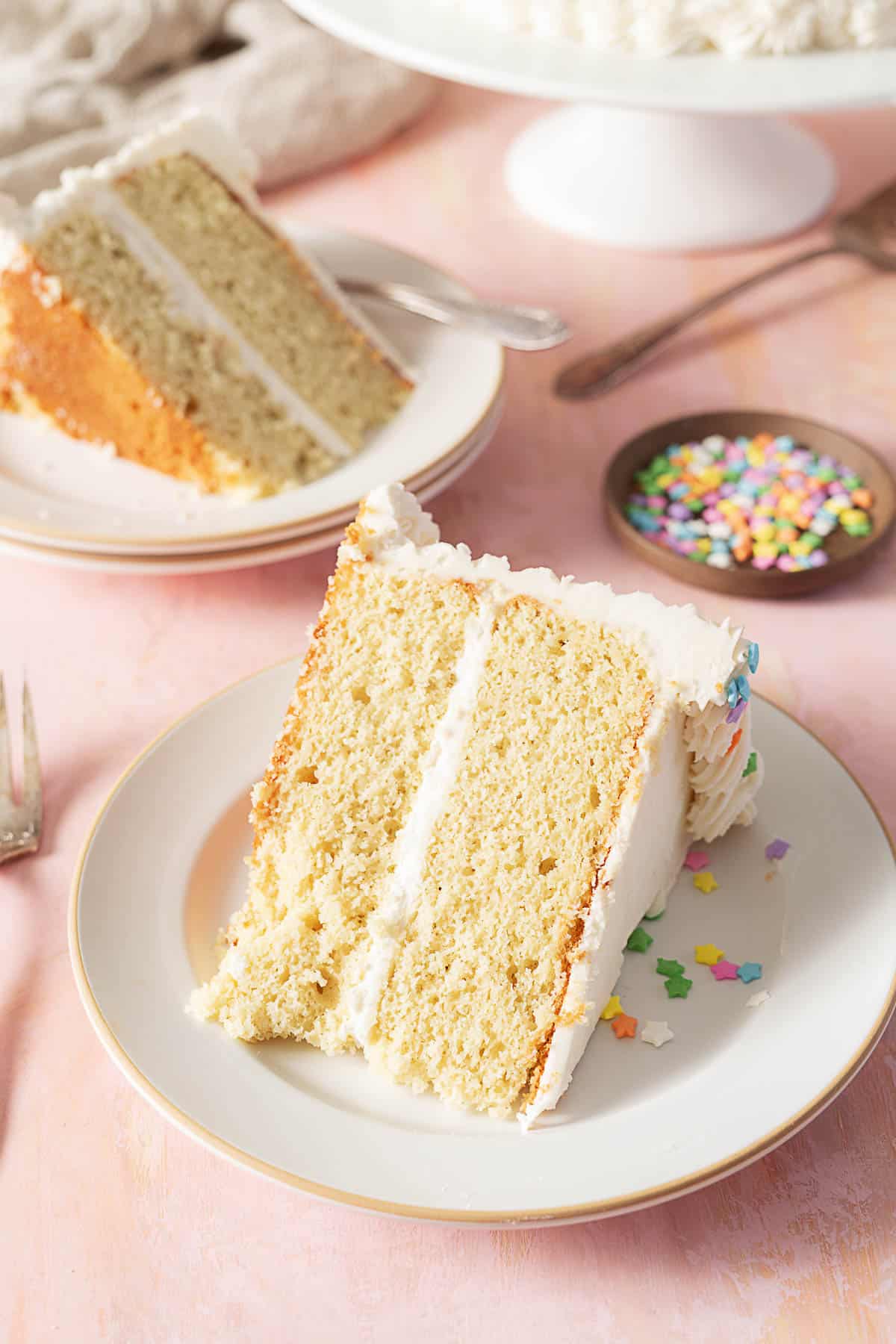 A slice of two-layer vanilla cake with white frosting on a plate, next to a fork and a small dish of colorful sprinkles.