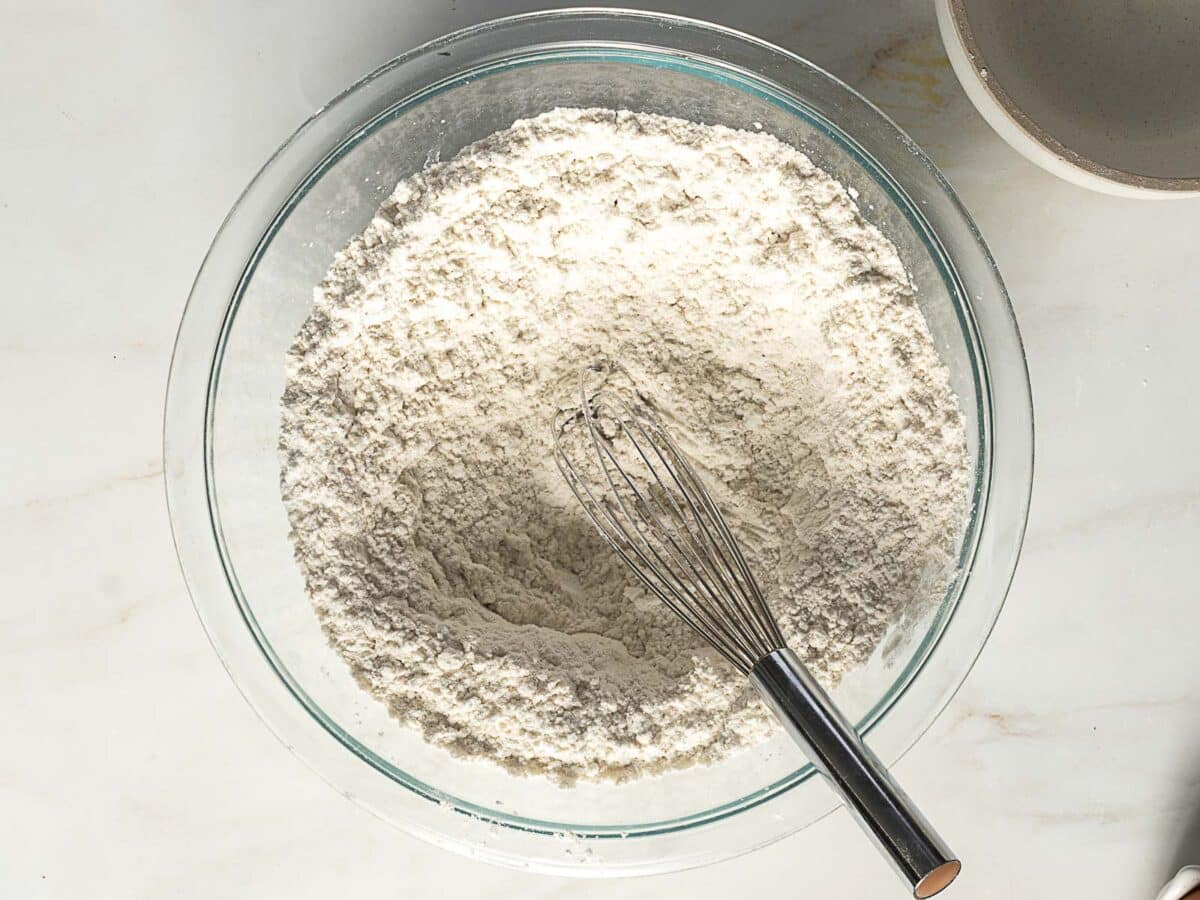 A glass bowl filled with flour mixture and a metal whisk placed on top, set on a light-colored countertop.