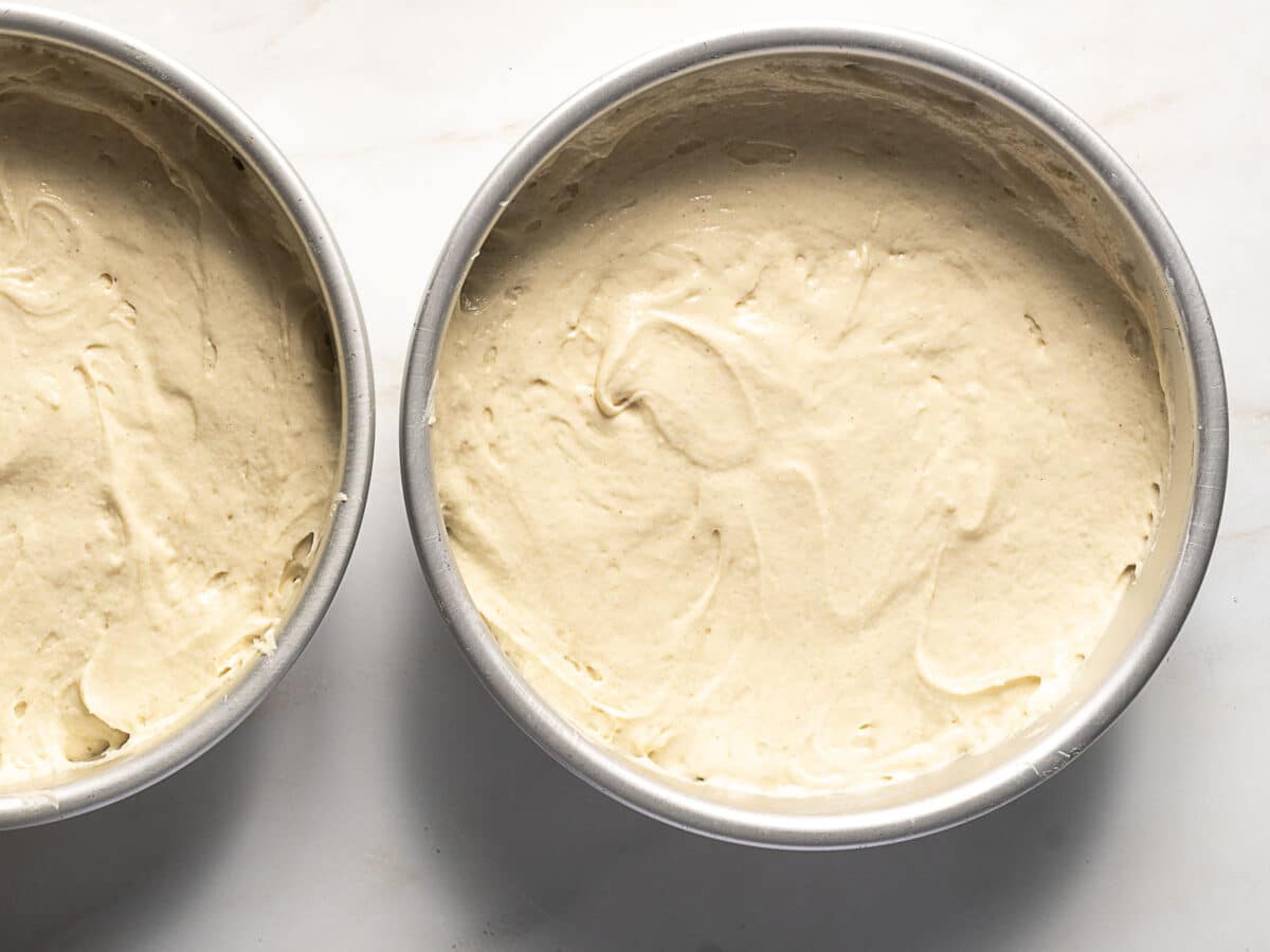 Two round metal cake pans filled with unbaked cake batter sitting on a light-colored surface.