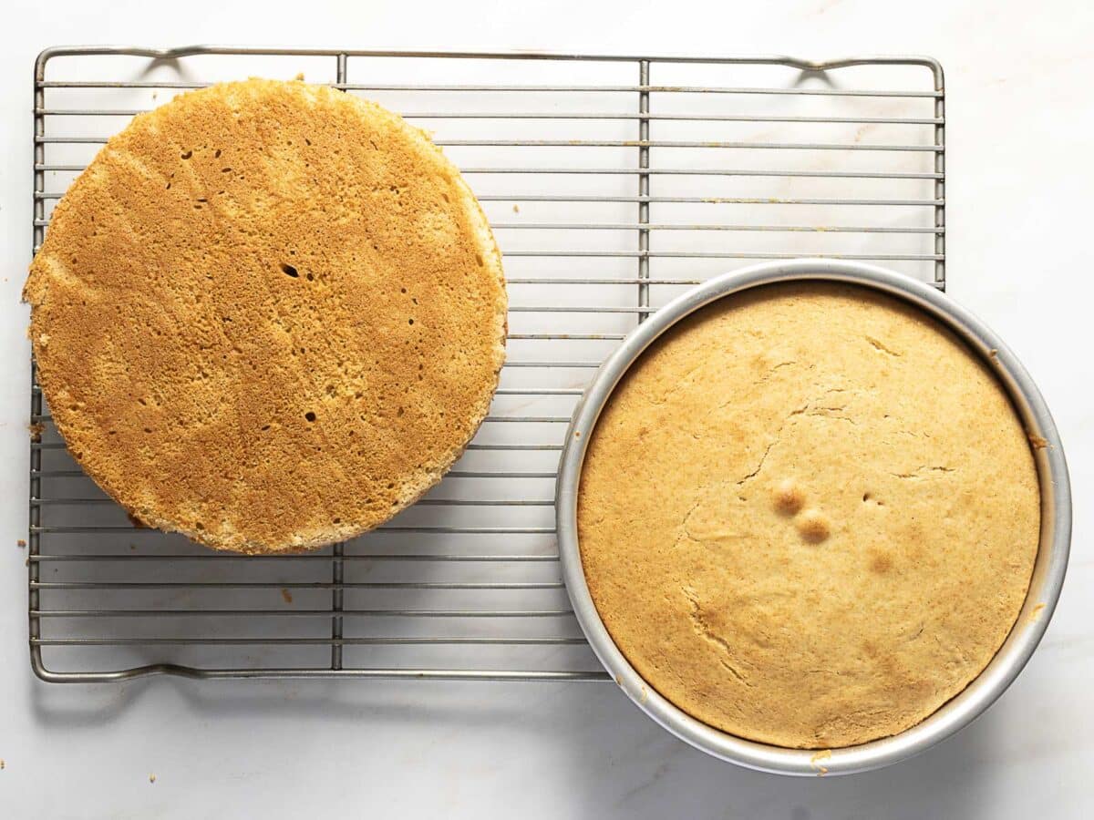 Two round sponge cakes, one on a cooling rack and one still in a baking pan, rest on a white surface.