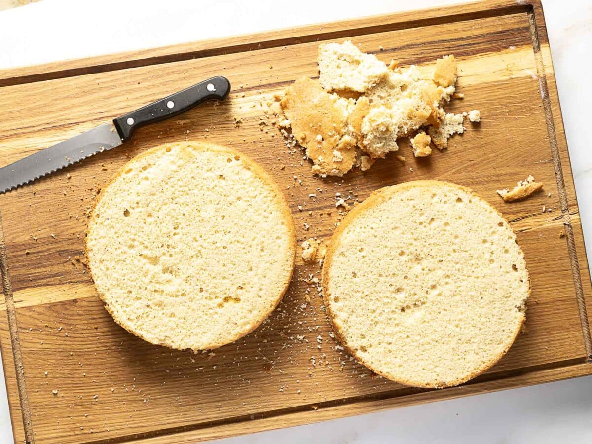 Two round cake layers, a serrated knife, and cake crumbs are on a wooden cutting board.