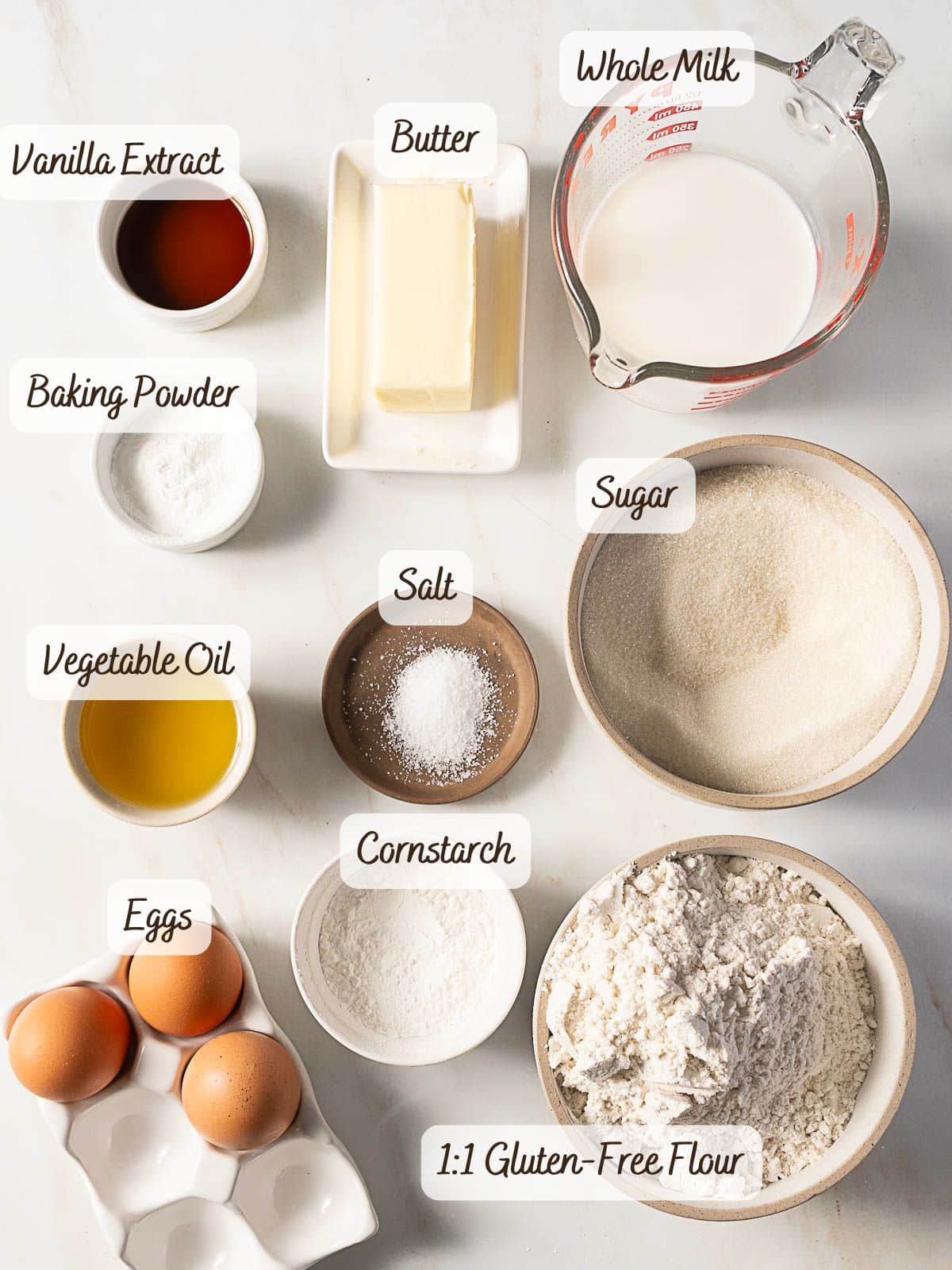 Top-down view of labeled baking ingredients in bowls and cups for a gluten free vanilla cake, including gluten-free flour, eggs, sugar, cornstarch, salt, baking powder, vanilla extract, vegetable oil, butter, and milk.