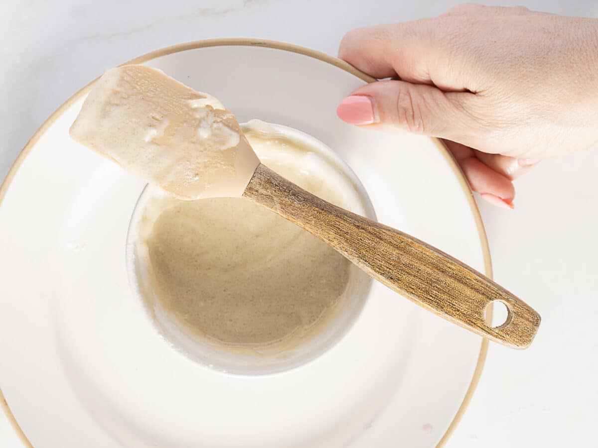A hand holding a plate with a small bowl of beige batter and a wooden spatula resting across the bowl.