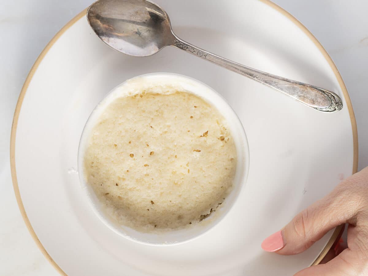 A small round dish of baked custard on a white plate with a silver spoon beside it and a hand holding the plate.