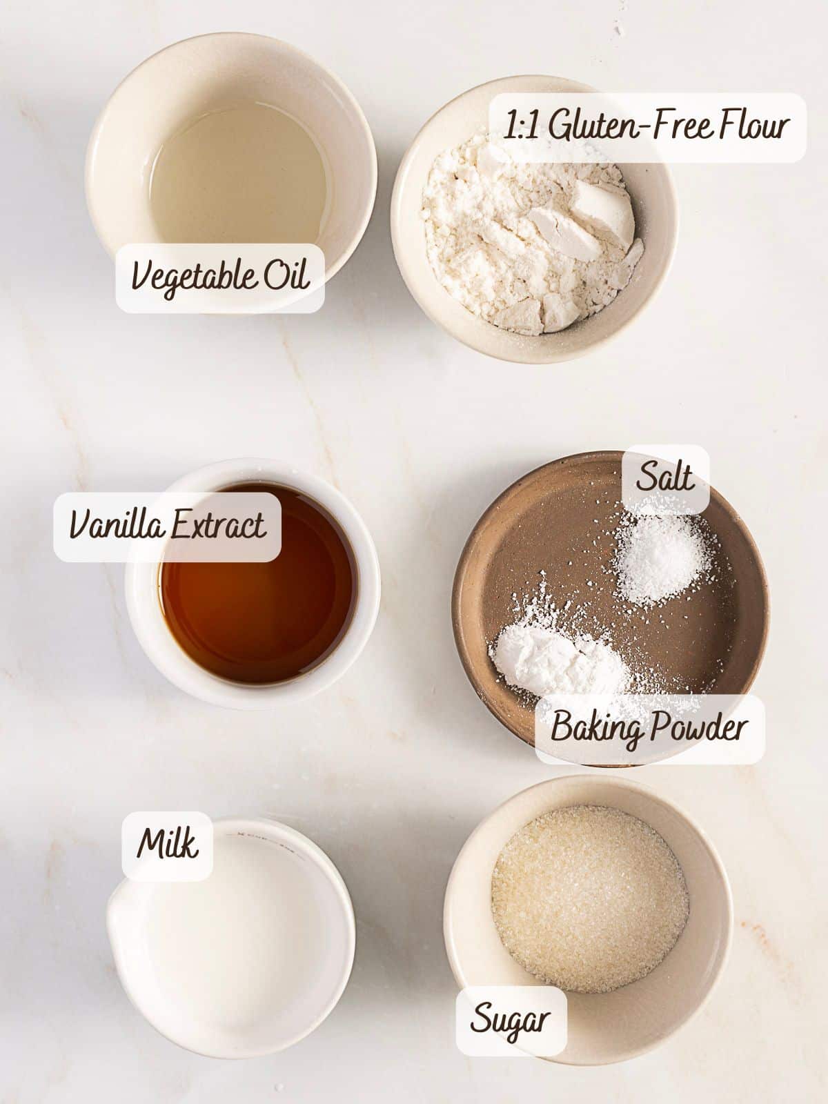 Six small bowls containing vegetable oil, gluten-free flour, vanilla extract, salt, baking powder, milk, and sugar are arranged on a white surface. Labels identify each ingredient.