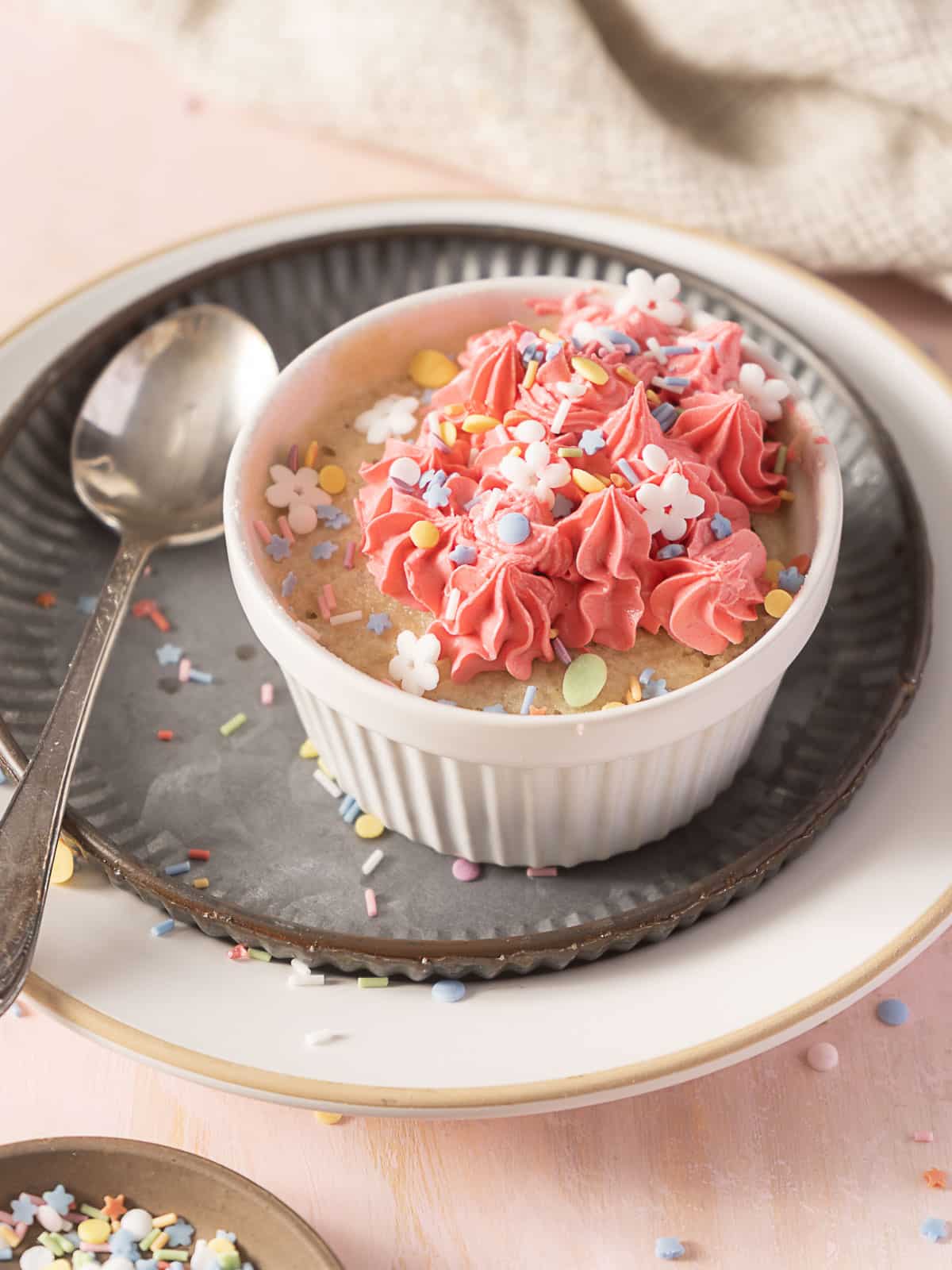 A small white ramekin of cake topped with pink frosting, colorful sprinkles, and flower-shaped decorations, placed on stacked plates with a spoon beside it.