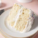 A slice of white layer cake with fluffy white frosting on a white plate, showing a light and airy texture.