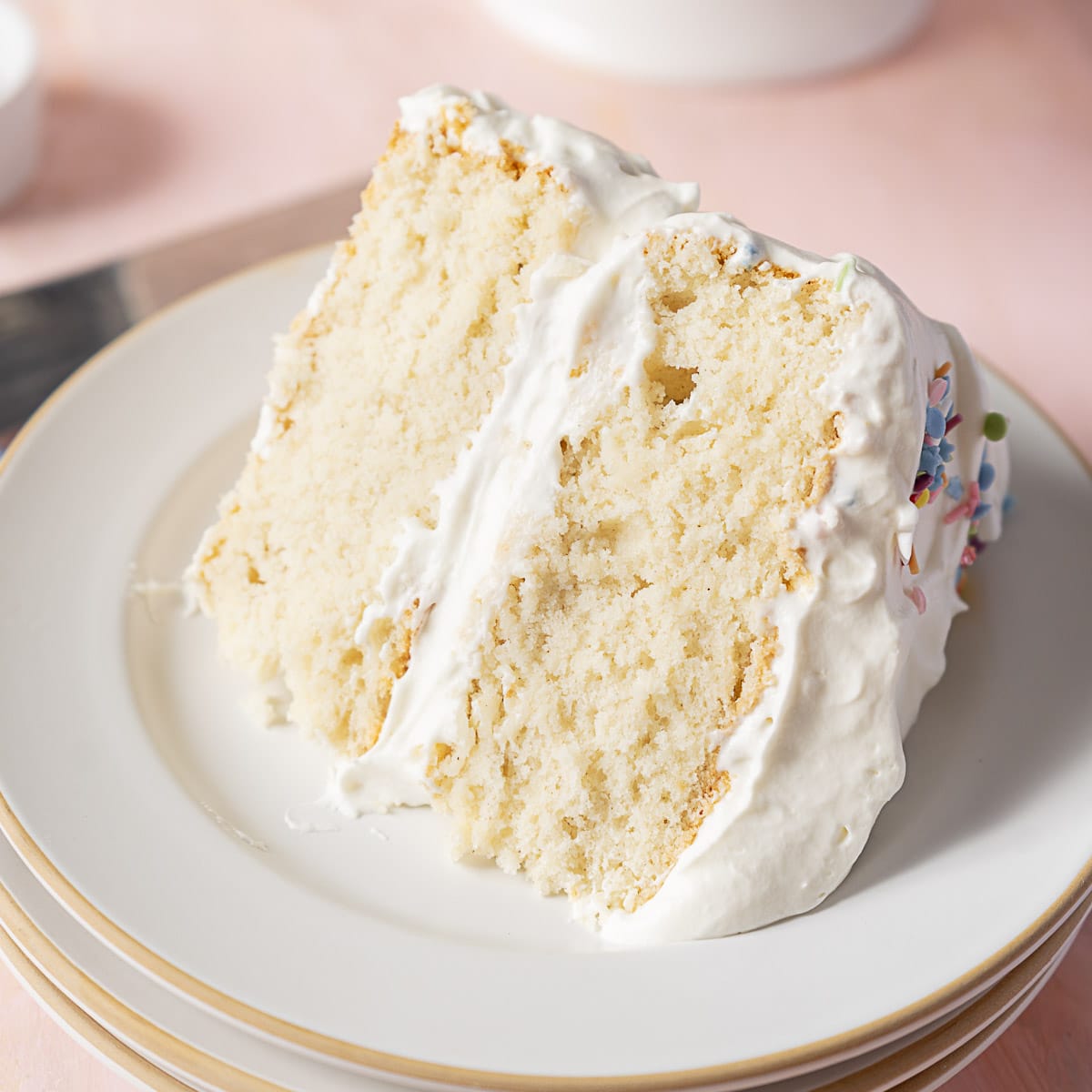 A slice of white layer cake with fluffy white frosting on a white plate, showing a light and airy texture.