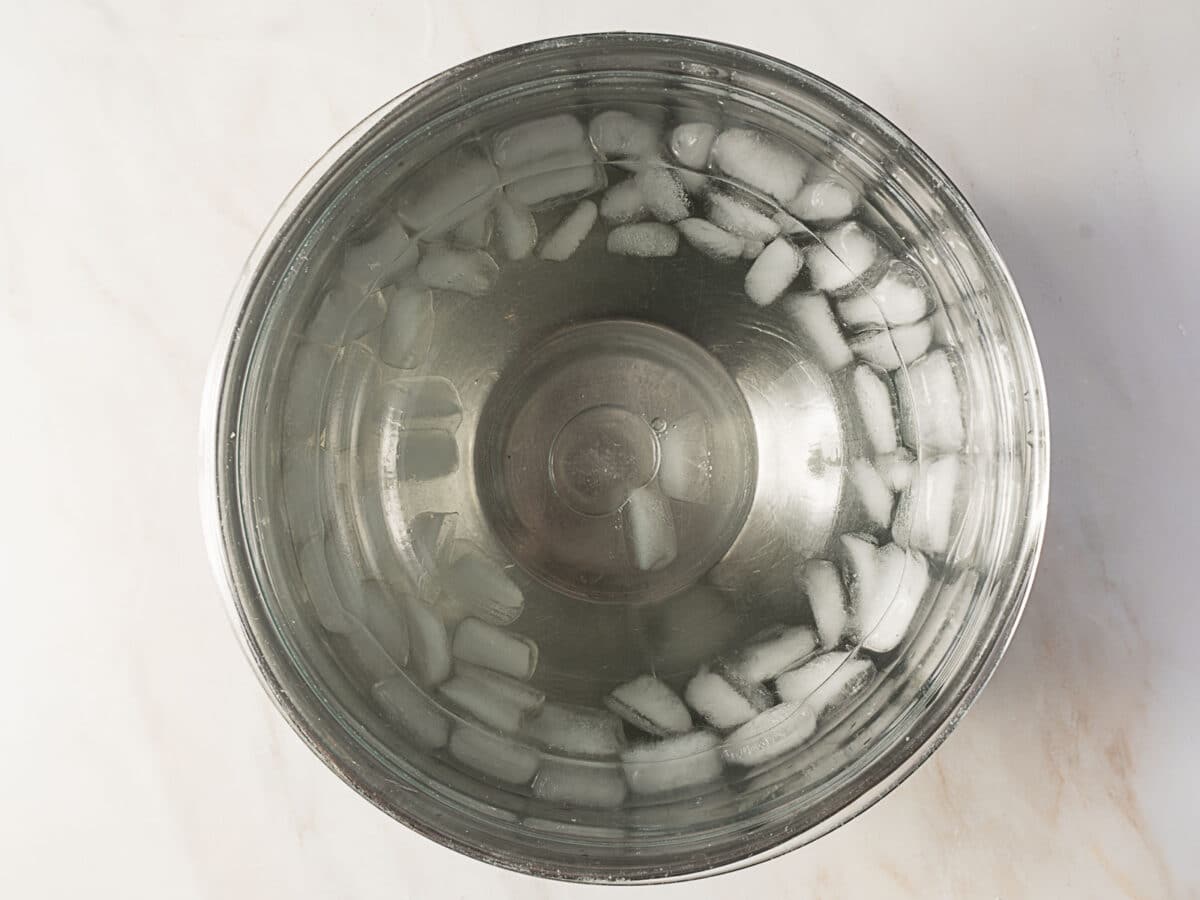 A stainless steel bowl filled with water and ice cubes, viewed from above on a light-colored surface, perfect for prepping buttermilk ice cream.