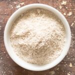 A white bowl filled with fine oat flour sits on a wooden surface with some flour and oat flakes scattered around.