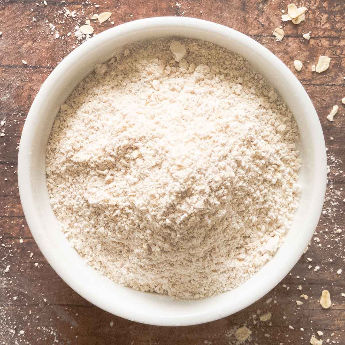 A white bowl filled with fine oat flour sits on a wooden surface with some flour and oat flakes scattered around.