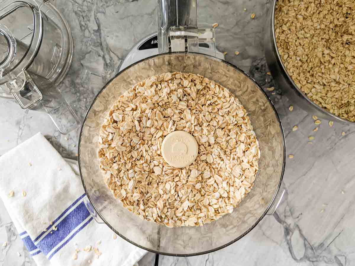 A food processor filled with rolled oats sits on a marble countertop next to a bowl of oats, a glass jar, and a white towel with a blue stripe.