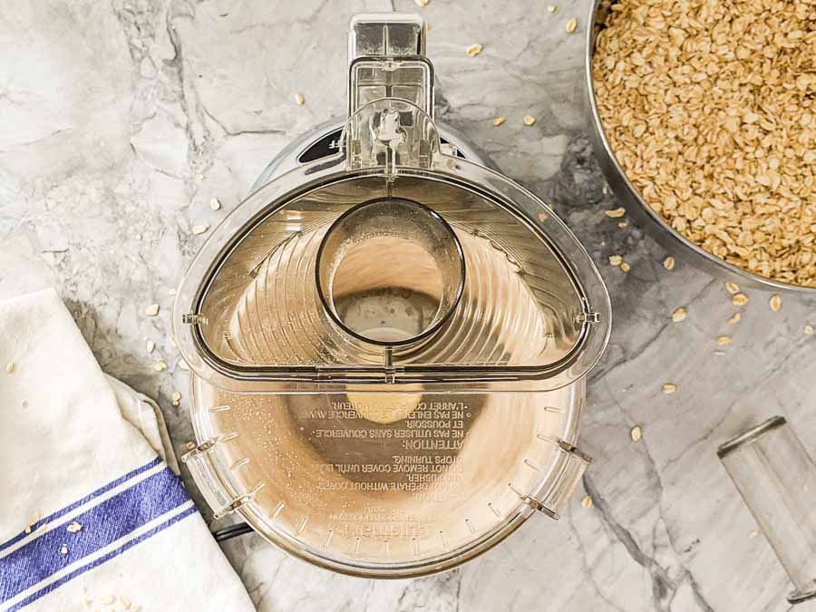 Top view of an empty food processor bowl on a marble counter, next to a metal bowl filled with oats and a white kitchen towel with a blue stripe.