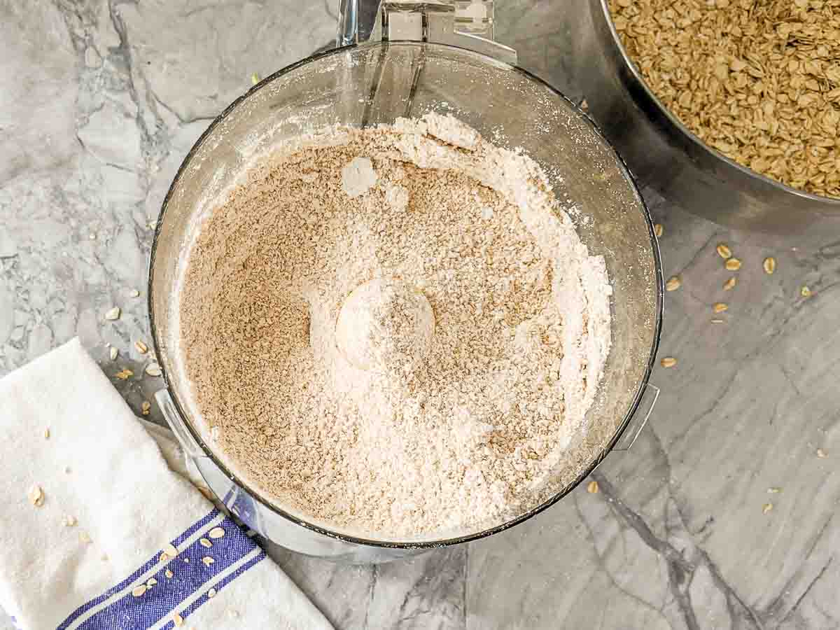 A food processor contains freshly ground oat flour; whole oats are visible in a nearby metal bowl on a gray marble counter.