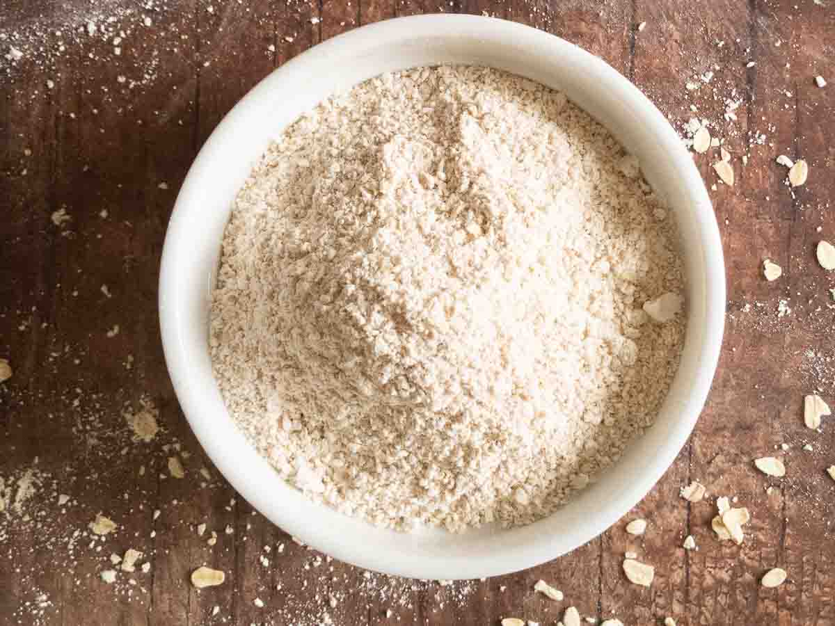 A white bowl filled with oat flour sits on a rustic wooden surface, with some flour and oat flakes scattered around.