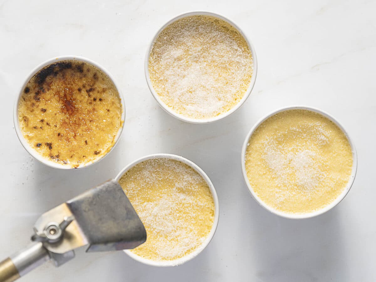 Four ramekins of crème brûlée are shown; one has a caramelized sugar top, while the others have uncooked sugar. A torch is visible near the ramekin with the caramelized top.