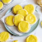 A plate of round lemon cookies with bright yellow frosting, arranged on parchment paper with additional cookies on the table and a cooling rack nearby.