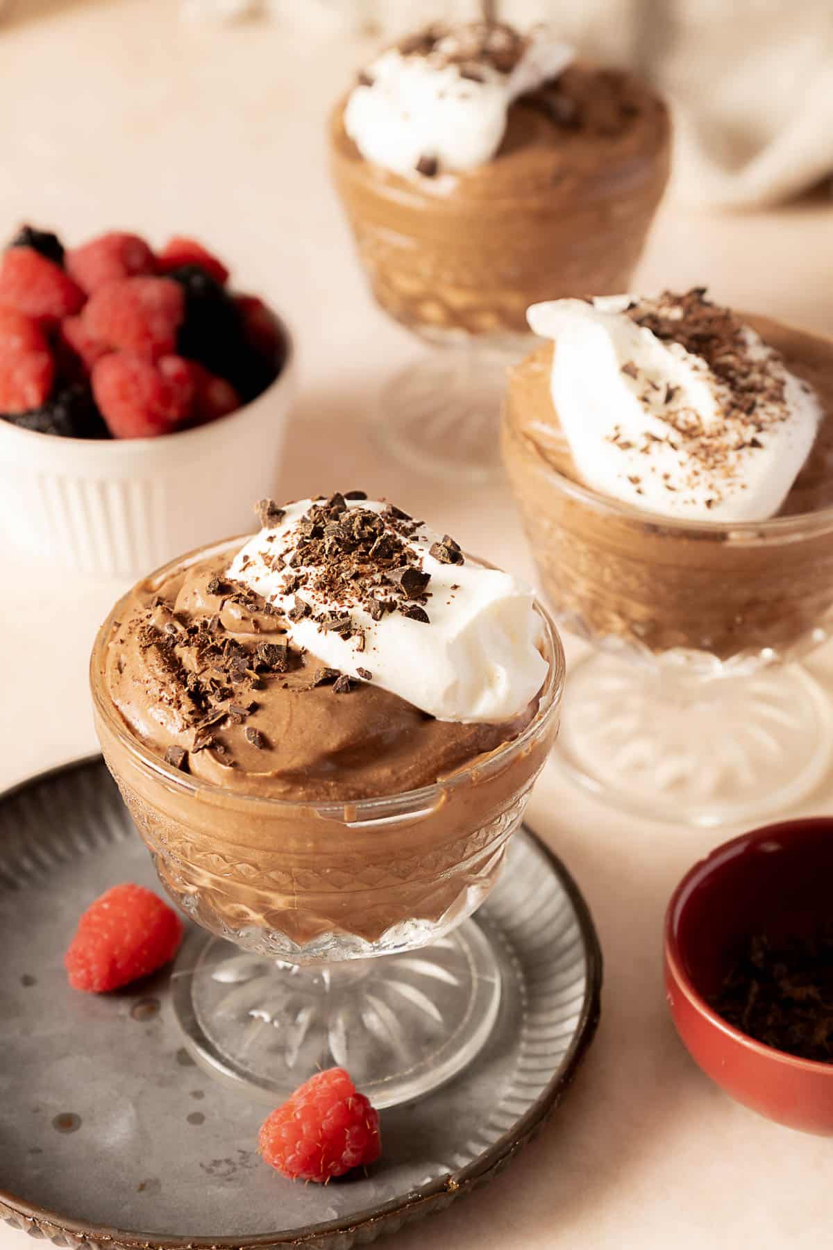 Three glass cups of chocolate mousse topped with whipped cream and chocolate shavings, with fresh raspberries and blackberries in the background.