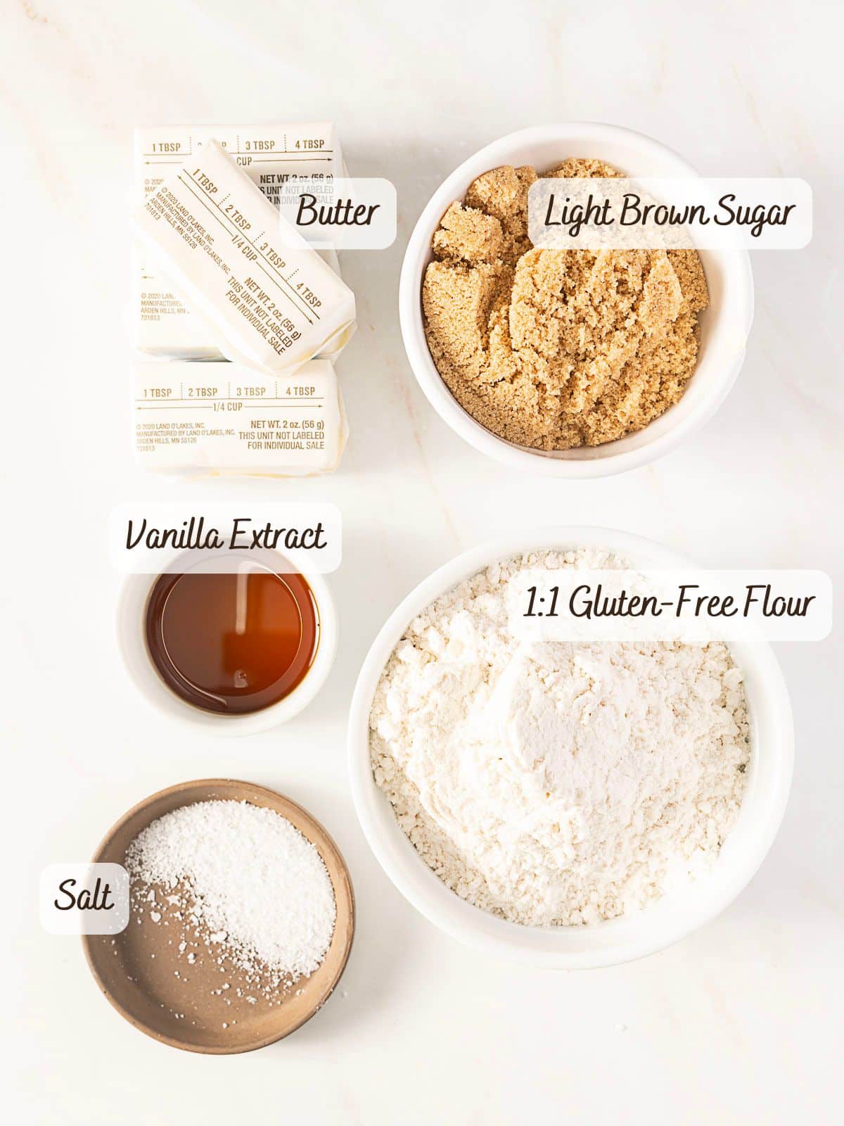 Five labeled ingredients for gluten free shortbread cookies: sticks of butter, a bowl of light brown sugar, a bowl of 1:1 gluten-free flour, a small bowl of salt, and a small bowl of vanilla extract on a white surface.