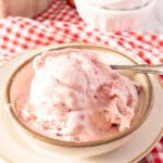 A bowl of strawberry ice cream with a spoon rests on a plate, placed on a red and white checkered cloth.
