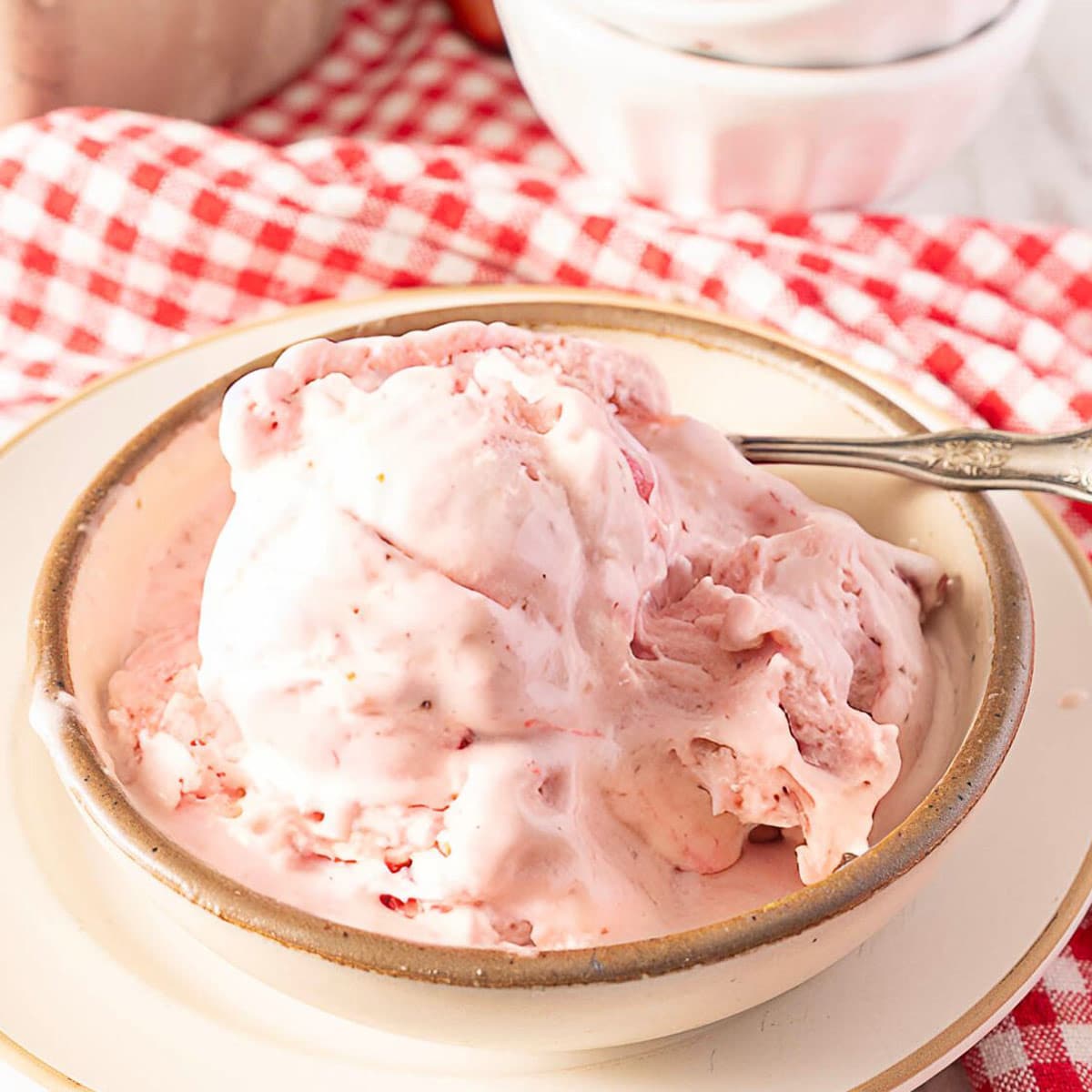 A bowl of strawberry ice cream with a spoon rests on a plate, placed on a red and white checkered cloth.