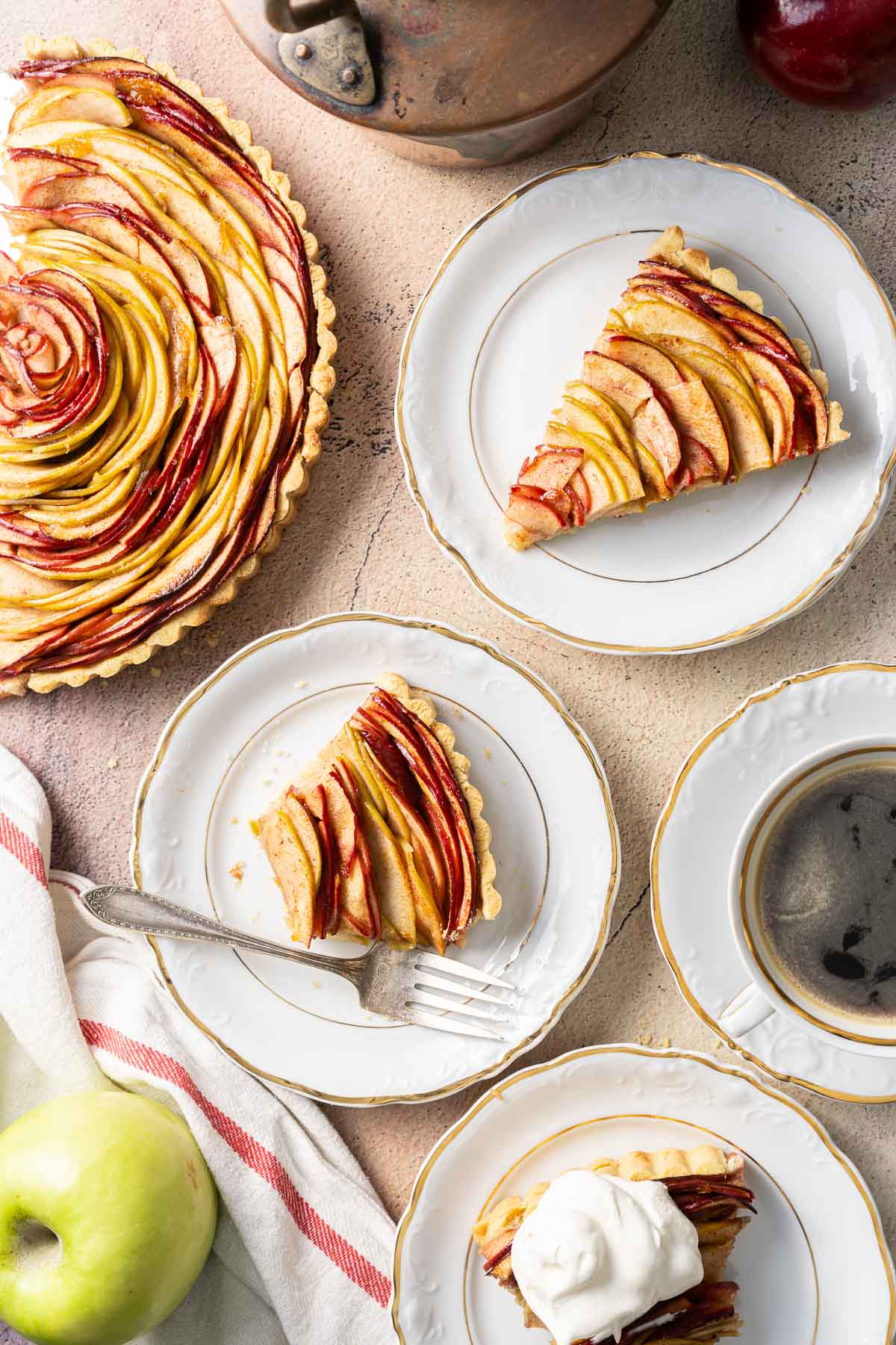 Sliced apple tart with three plated servings, one topped with whipped cream, alongside a cup of coffee and a green apple on a light tabletop.