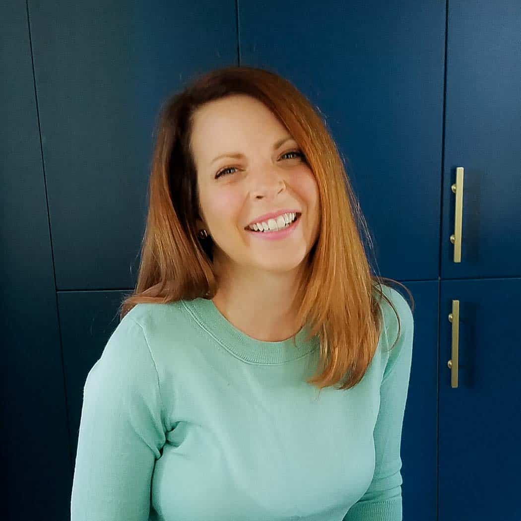 A woman with straight, shoulder-length brown hair smiles while wearing a light blue sweater, standing in front of dark blue cabinets—ready to share her favorite easy, gluten free desserts.