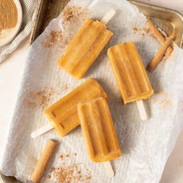 Four orange popsicles are arranged on a parchment-lined baking tray, with cinnamon sticks and ground cinnamon sprinkled nearby.