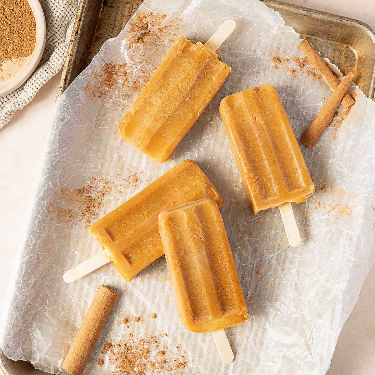 Four orange popsicles are arranged on a parchment-lined baking tray, with cinnamon sticks and ground cinnamon sprinkled nearby.