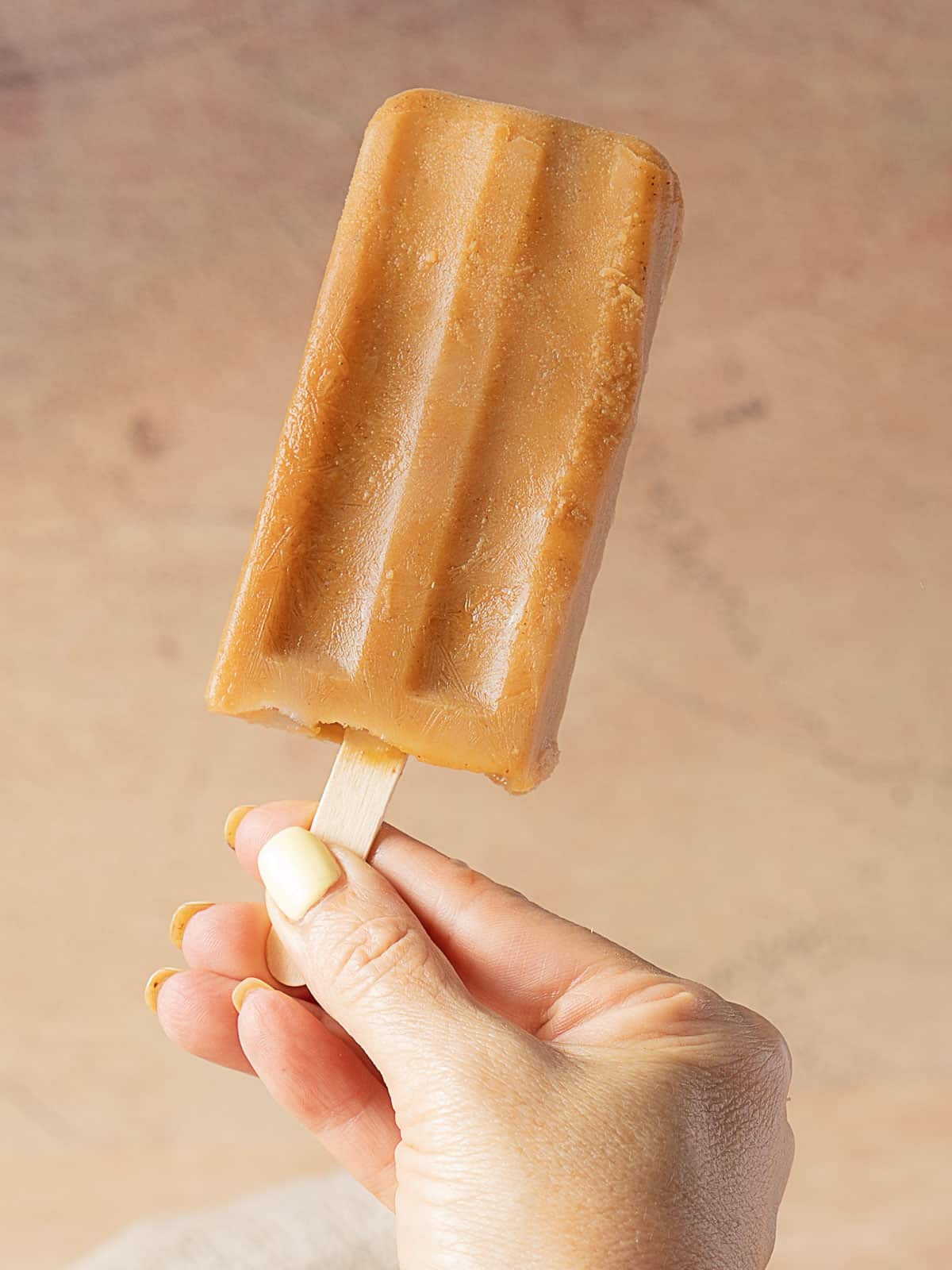 A hand holding a brown popsicle on a wooden stick against a neutral background.