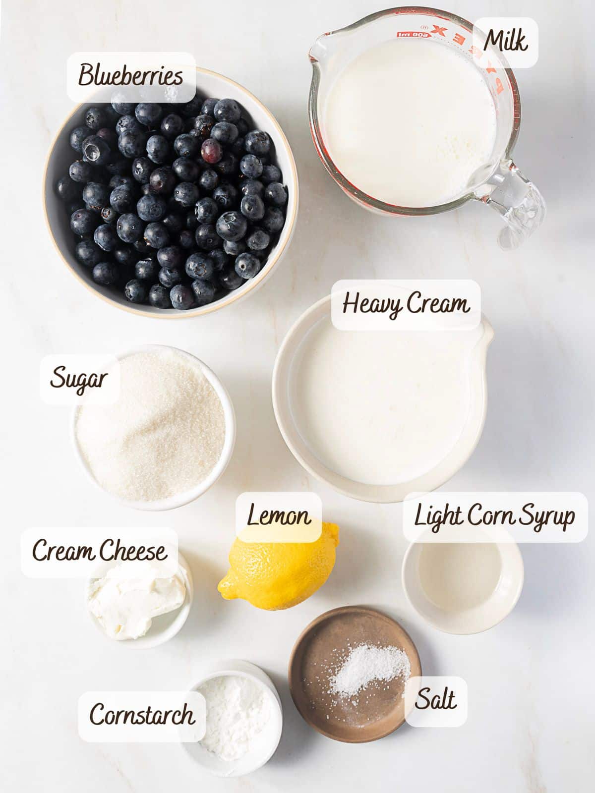 Overhead view of labeled ingredients for no eggs blueberry ice cream: blueberries, milk, heavy cream, sugar, cream cheese, lemon, cornstarch, light corn syrup, and salt arranged neatly on a white surface.