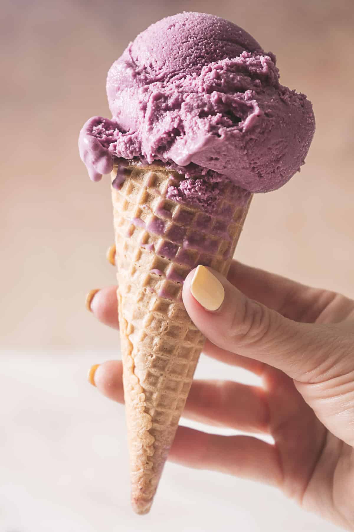 A hand with painted nails holds a waffle cone with two scoops of purple ice cream against a neutral background.