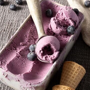 A metal scoop in a rectangular container of purple ice cream with blueberries on top, next to an empty waffle cone on a textured gray surface.