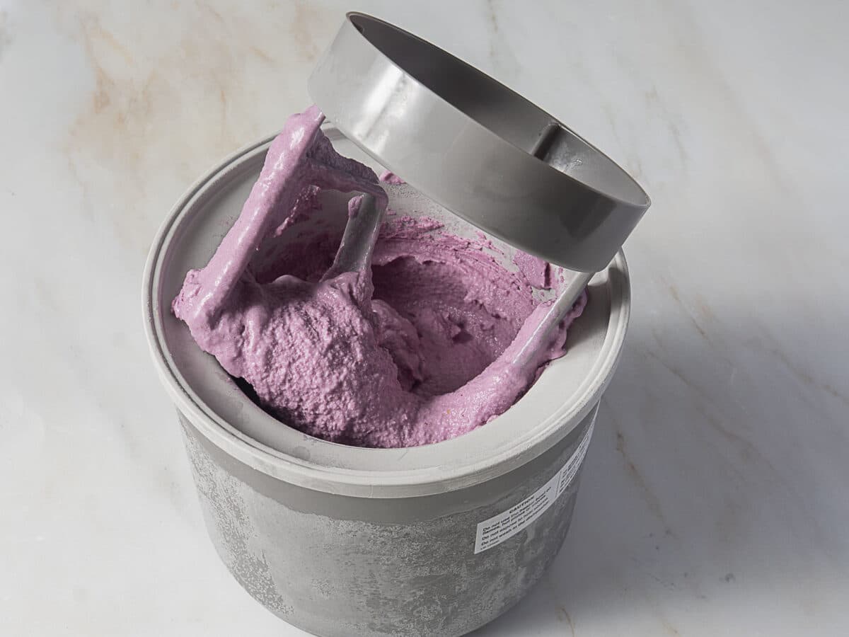 A container of purple ice cream with a mixing paddle attached to the lid, resting on a light marble surface.