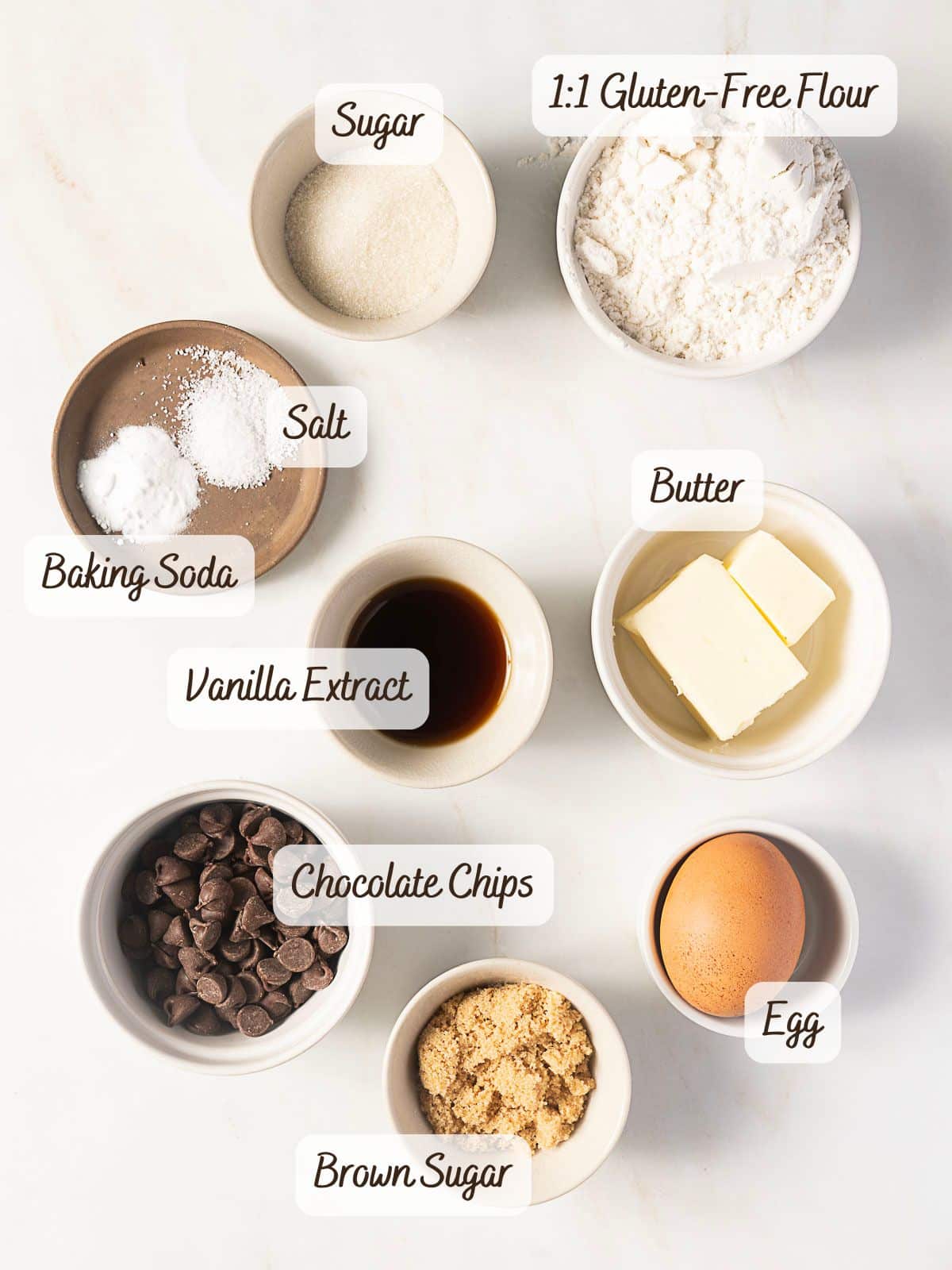 Assorted baking ingredients in bowls labeled as 1:1 gluten-free flour, sugar, salt, baking soda, butter, vanilla extract, chocolate chips, egg, and brown sugar—perfect for making Gluten Free Air Fryer Cookies.