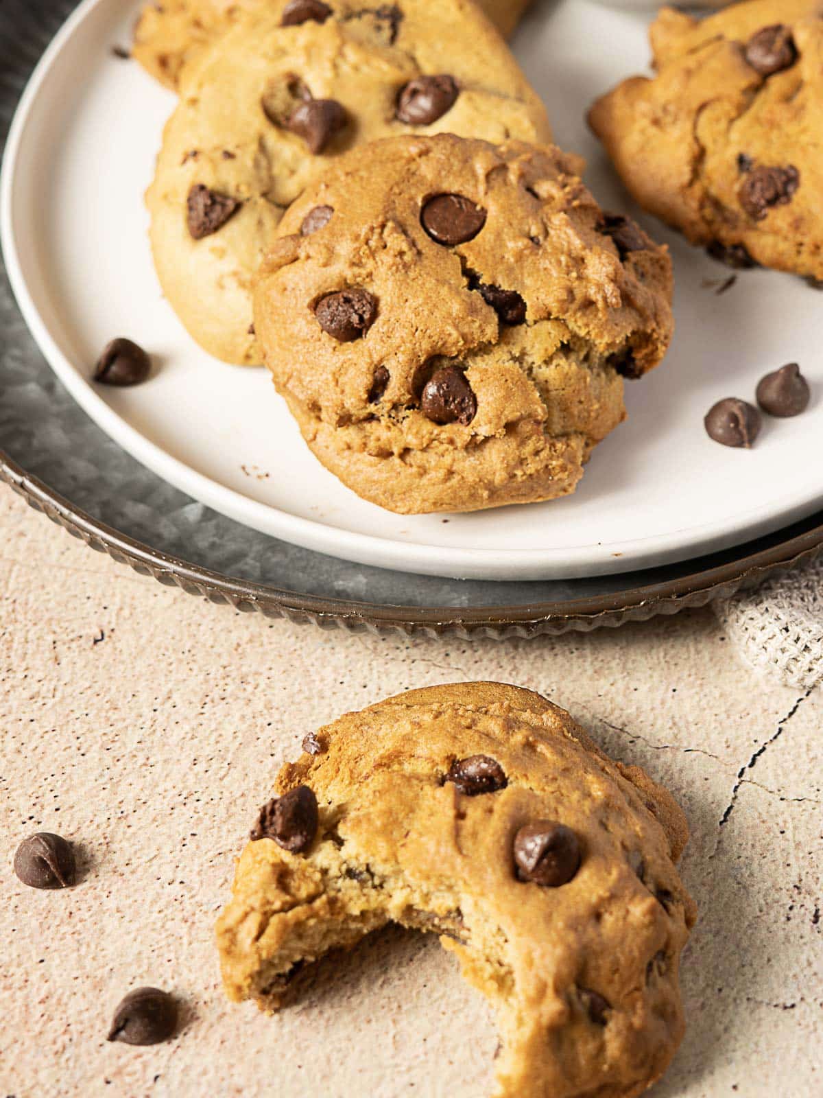 A plate with several chocolate chip cookies and a single cookie with a bite taken out of it on the textured surface nearby. Chocolate chips are scattered around.