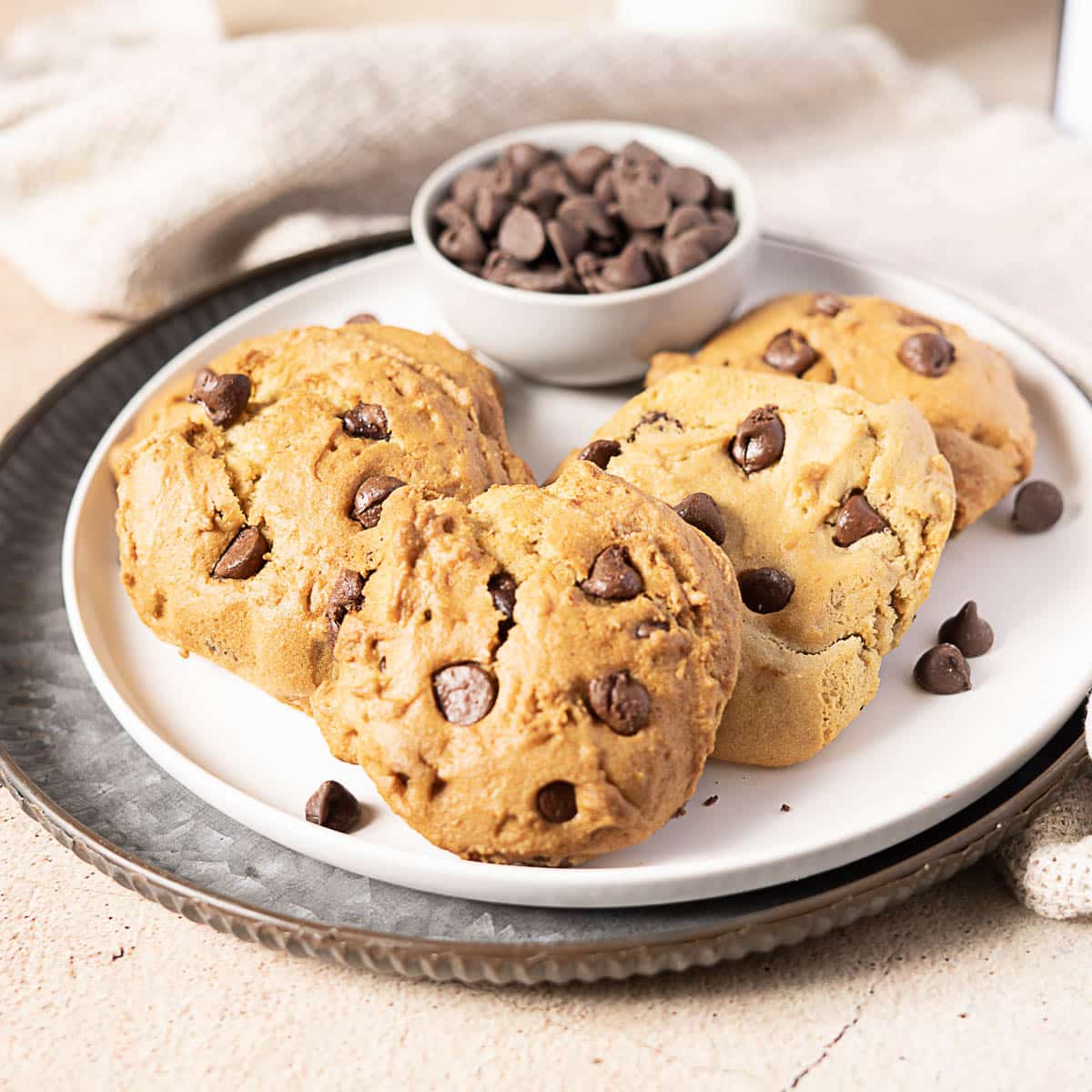 A plate with four chocolate chip cookies and a small bowl of chocolate chips, placed on a textured surface.